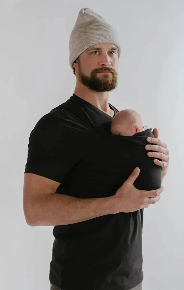 Black baby carrier sling T-shirt with short sleeves and front wrap, holding baby.