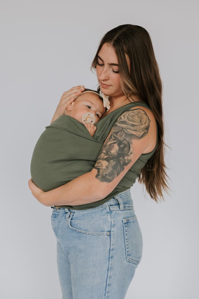 Olive green baby carrier sling T-shirt with front wrap panel, worn while holding baby.